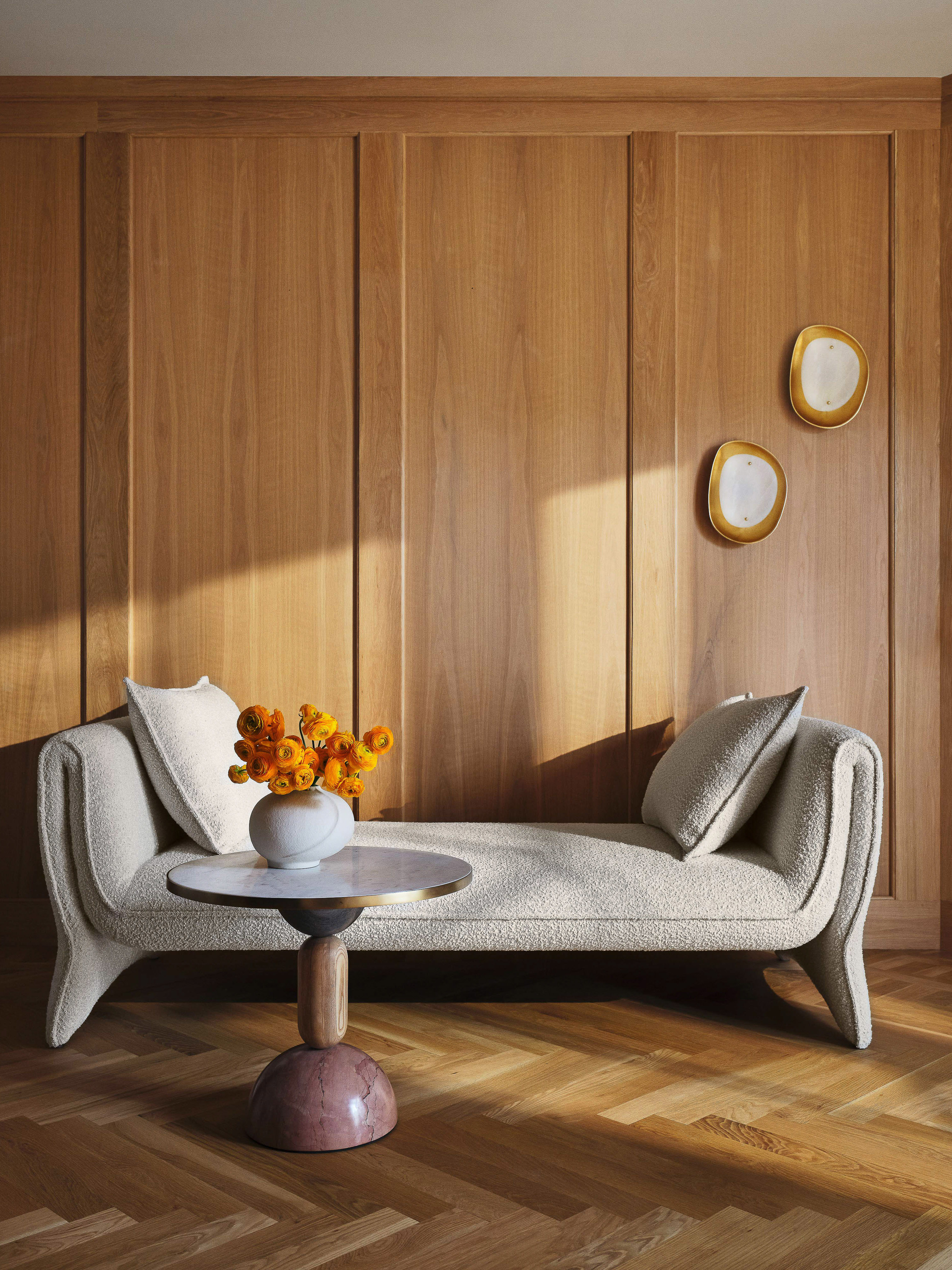 Interior shot of a daybed with pillows, a marble-topped side table with flowers, set against a wood-paneled wall and herringbone floor.