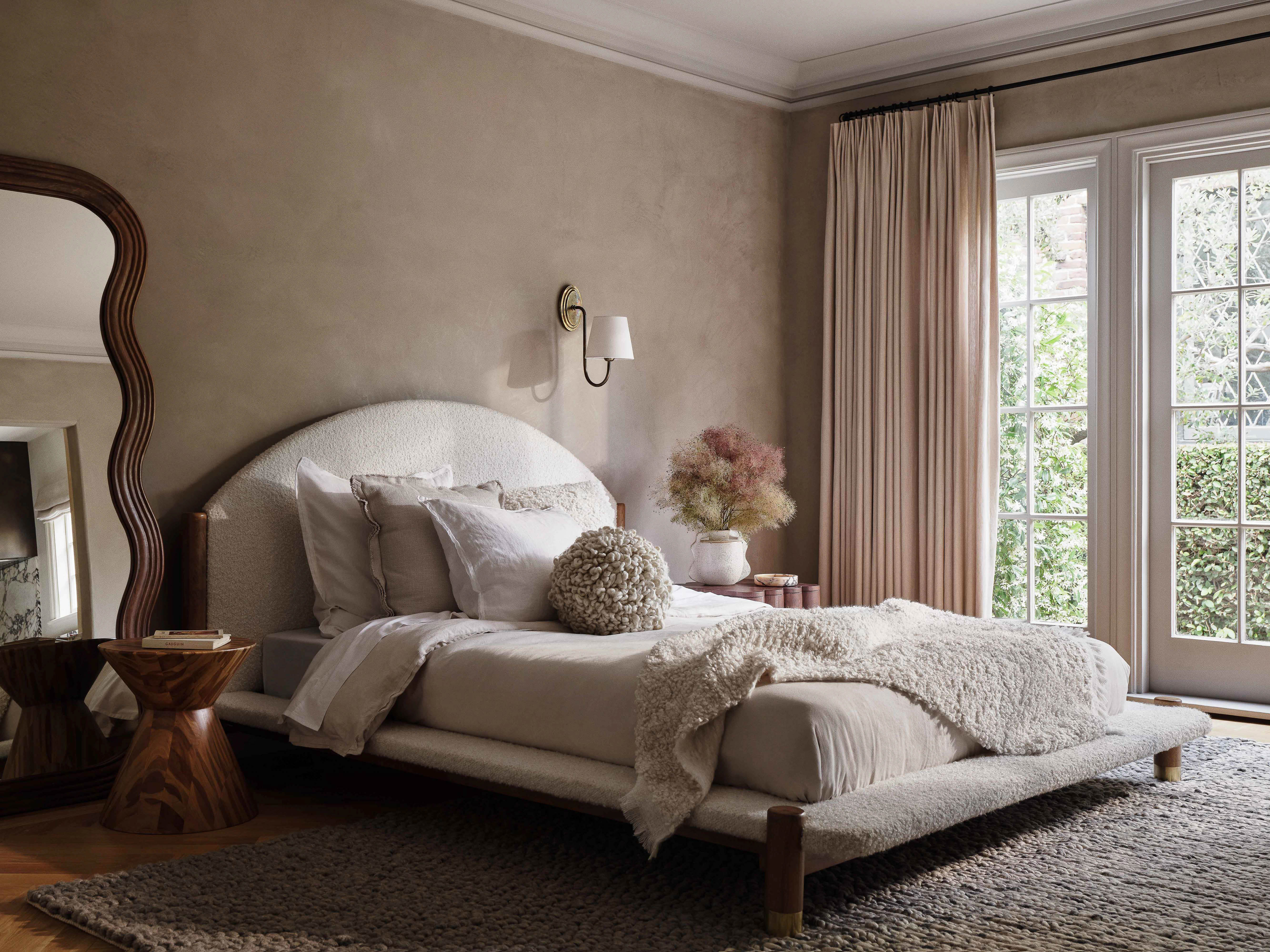 Bedroom interior featuring a bed with white linens and headboard, a wooden side table, a decorative mirror, and natural light from windows.