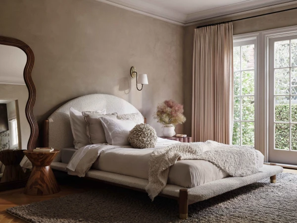 Bedroom interior featuring a bed with white linens and headboard, a wooden side table, a decorative mirror, and natural light from windows.