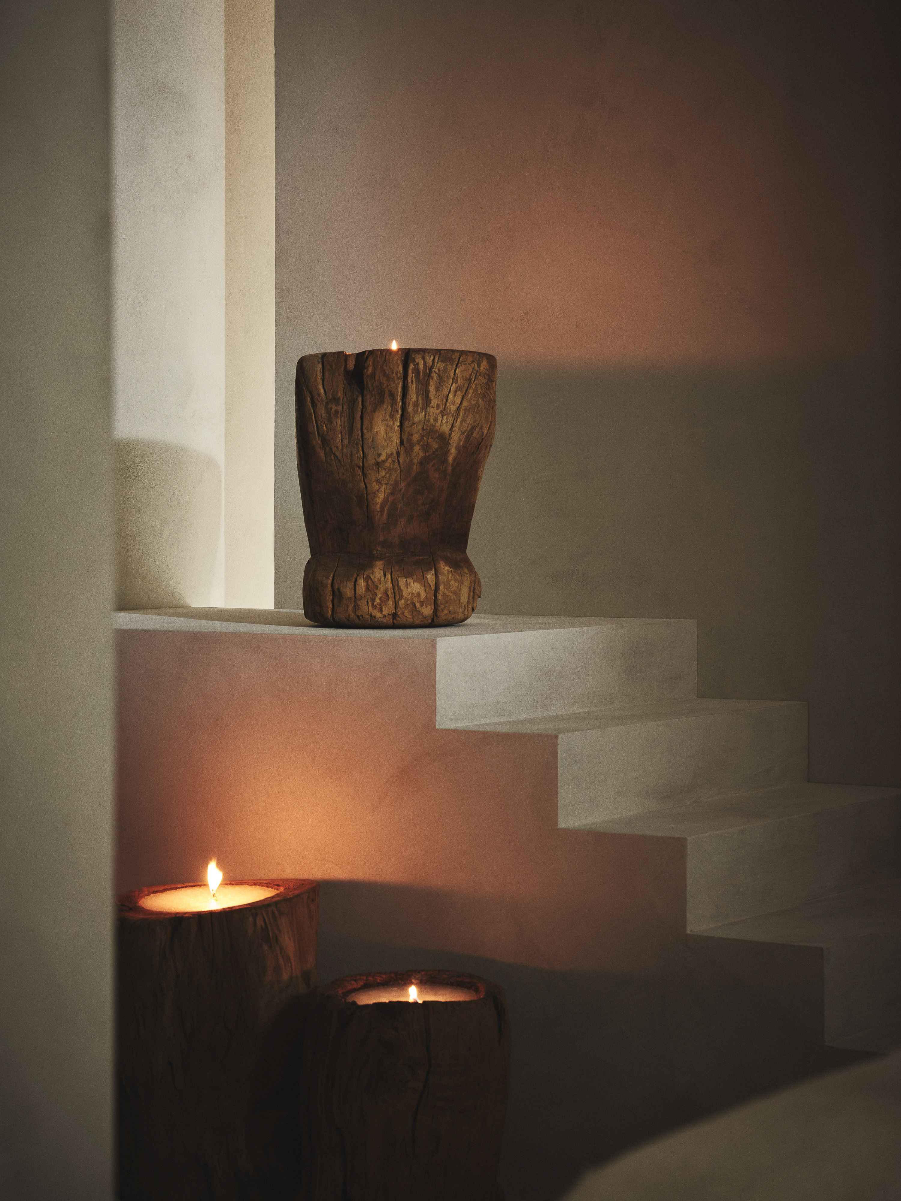Two wooden candle holders with lit candles sit on white stairs, with a third candle holder on top step, in a neutral interior space.