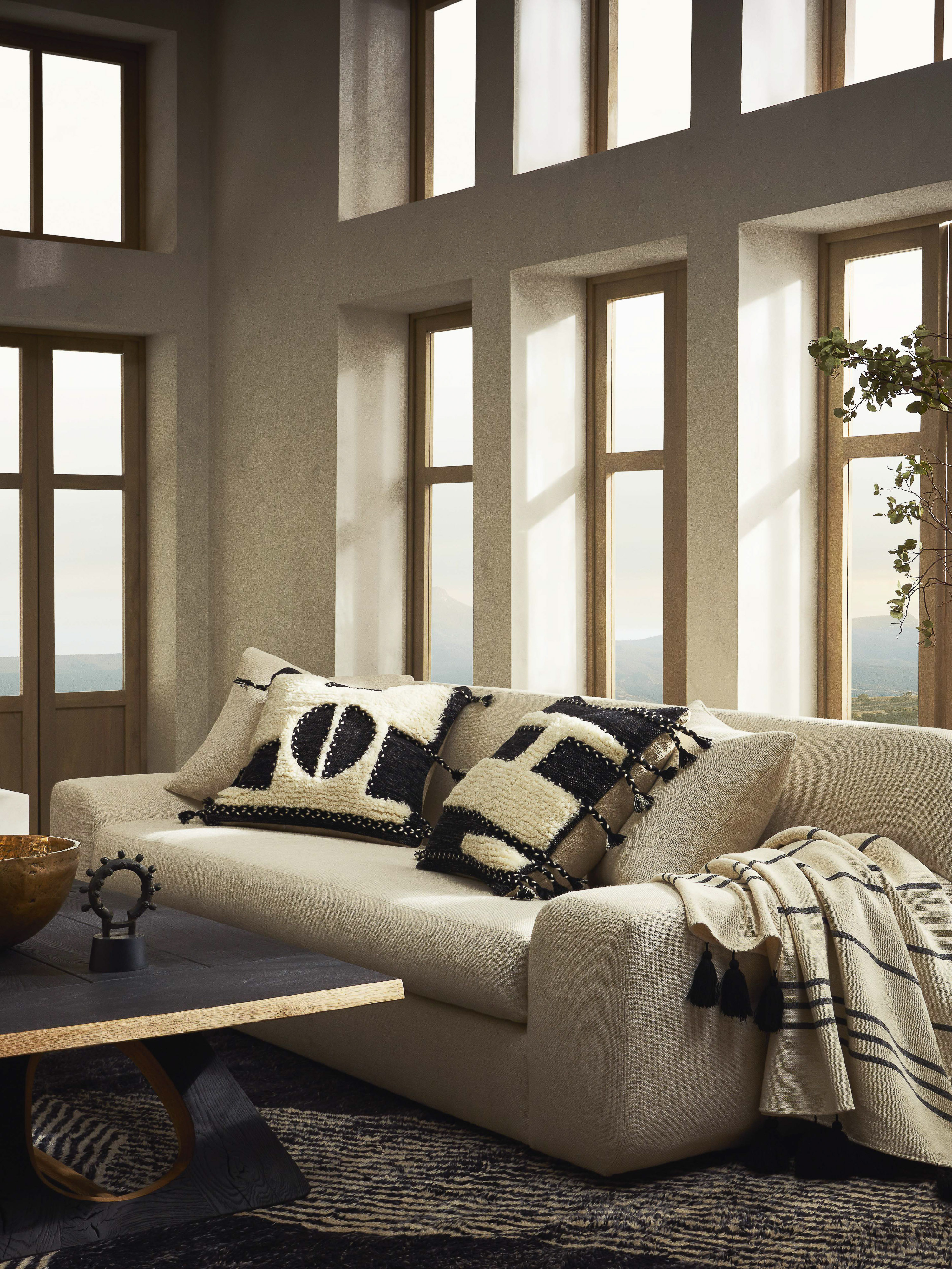 Living room interior featuring a cream sofa with decorative cushions, a dark coffee table, and natural light from large windows.