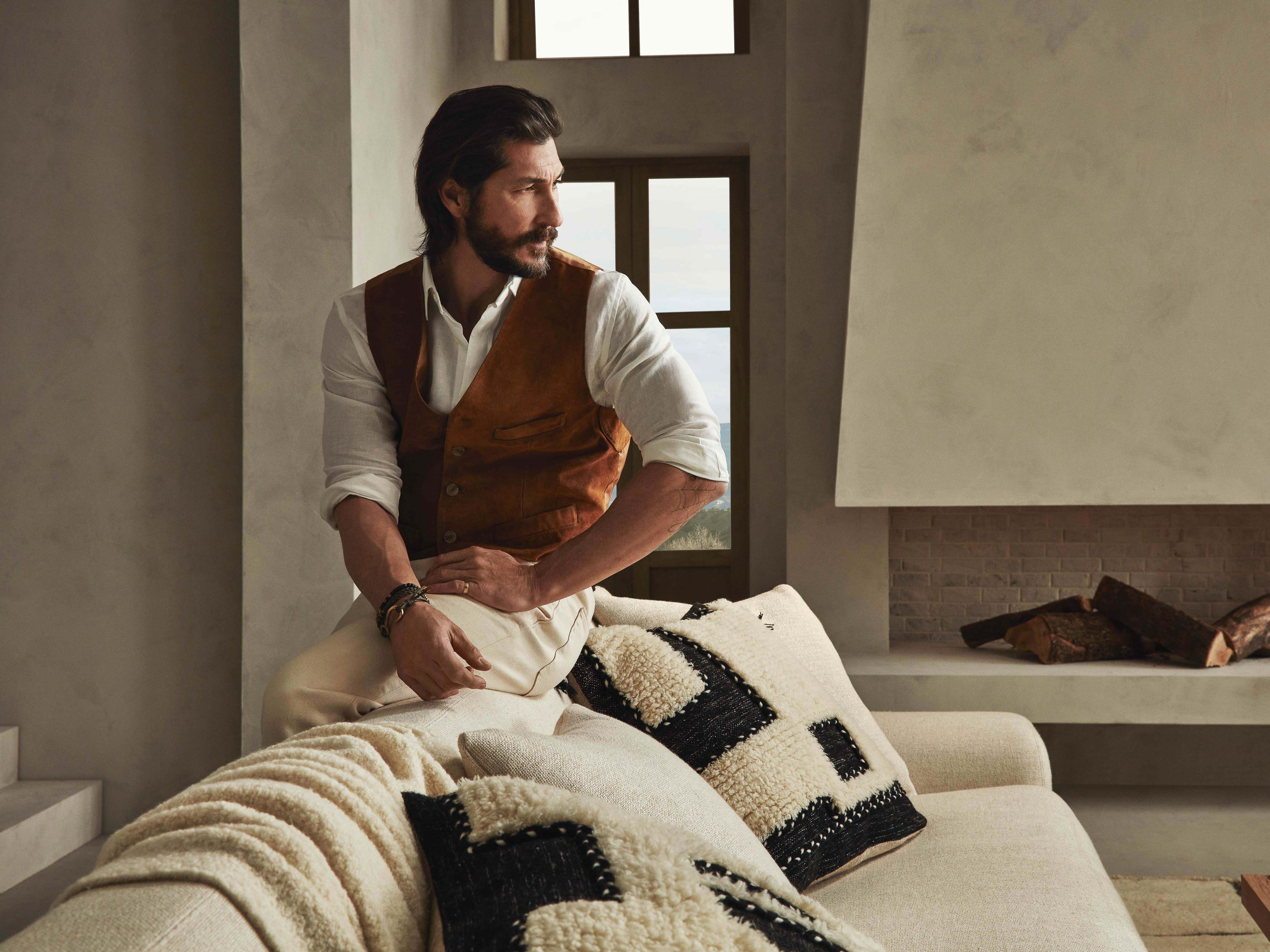 Man in a brown vest sits on a sofa in an indoor setting with neutral tones, textured pillows, and soft natural lighting.