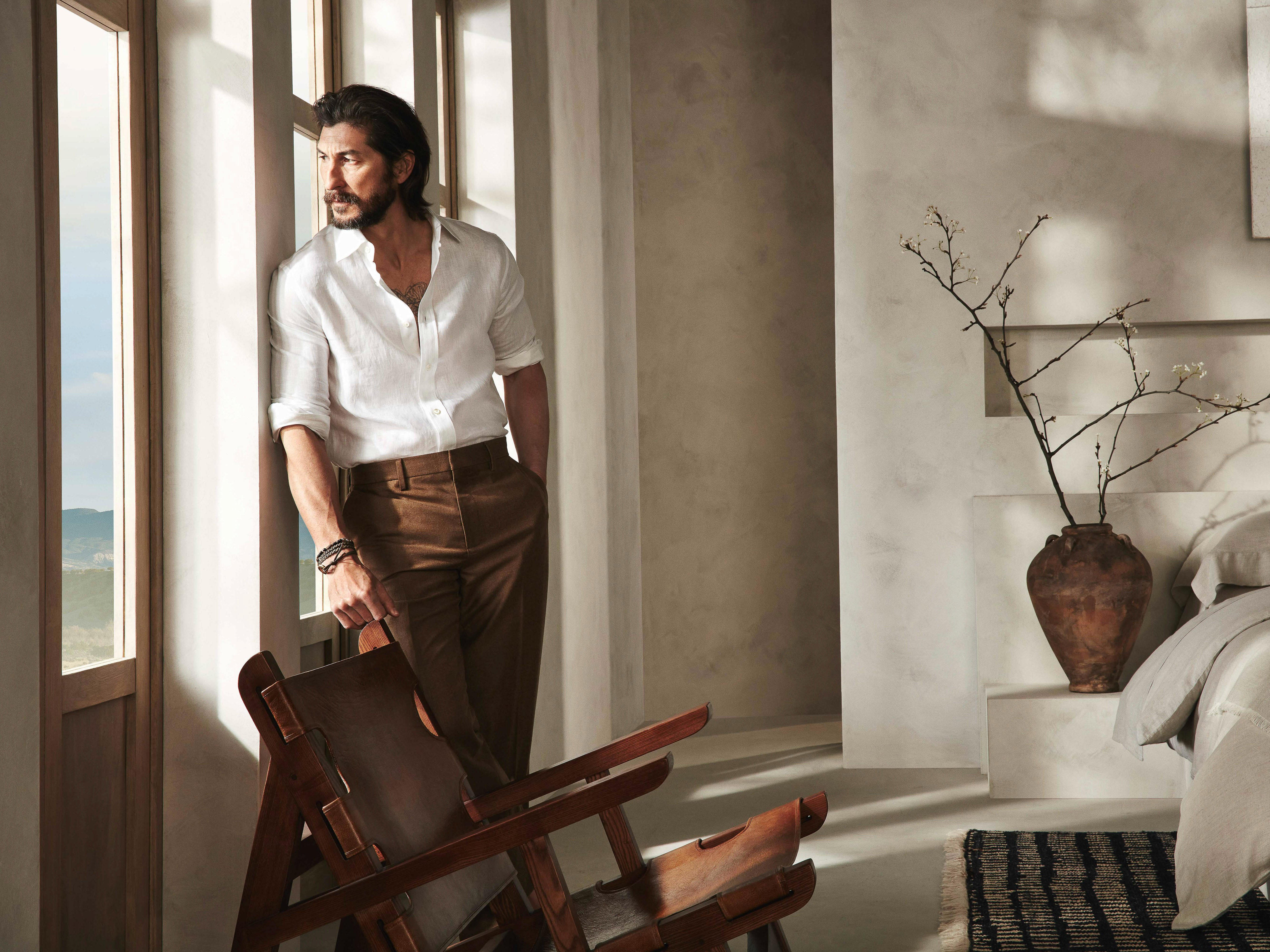 Adult male in a white linen shirt and brown trousers leans against a window in a minimally decorated room with earth-tone colors.