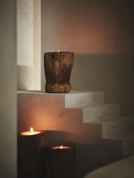 Three rustic carved wood candles glowing on and beside minimalist plaster steps in warm, moody lighting.