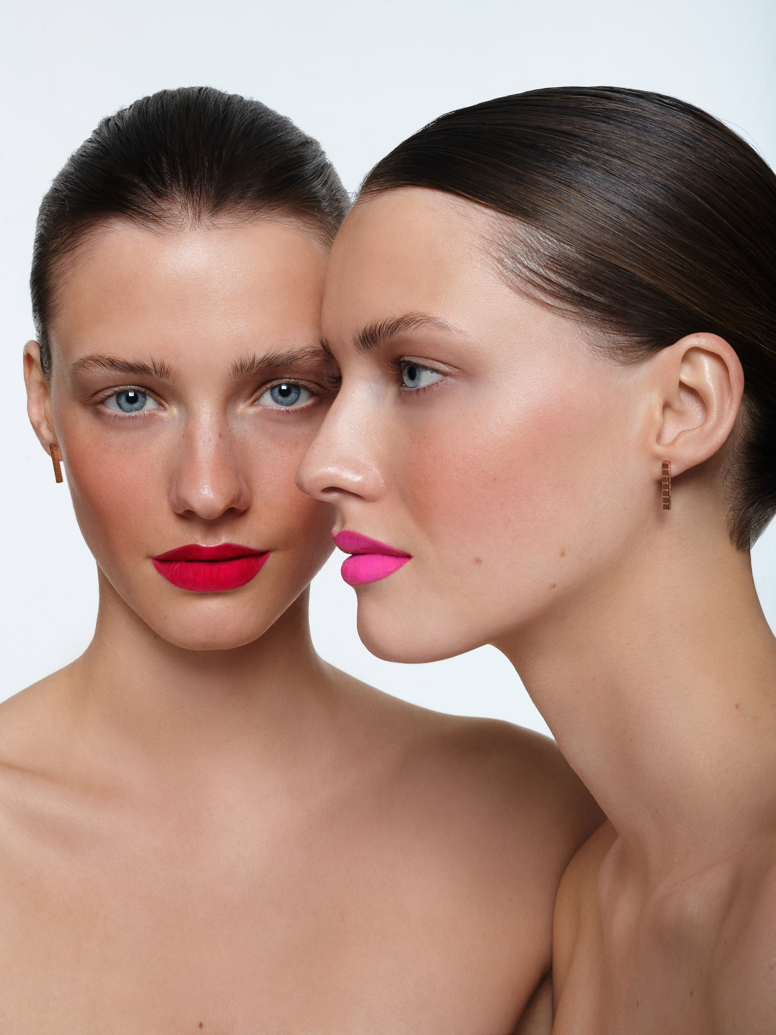 Two fair-skinned women with bold lipstick colors—red and pink—pose closely in a beauty shot with a clean, bright white backdrop.