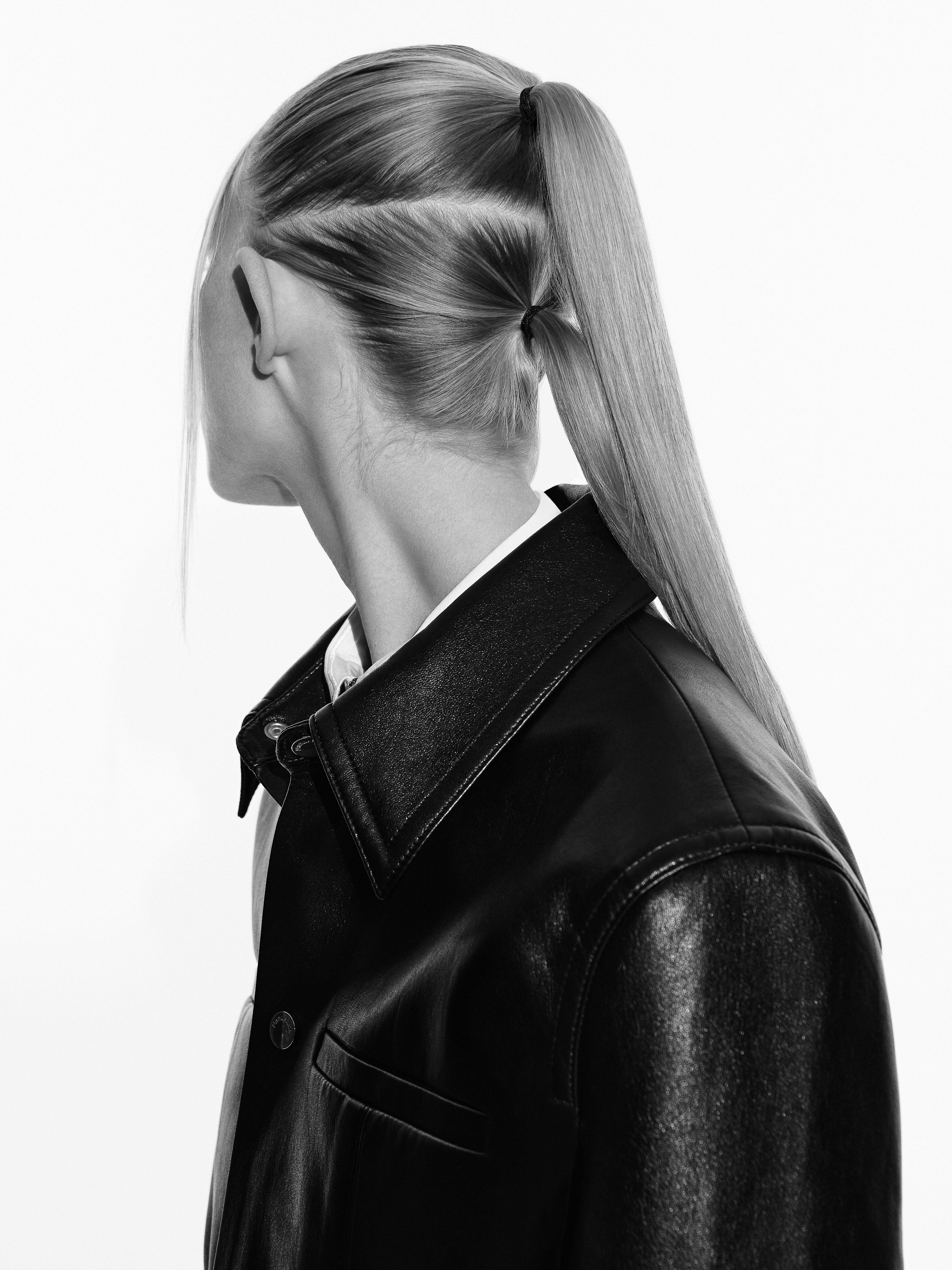 Black and white studio shot of a woman with a blonde ponytail wearing a black leather jacket on a white background.