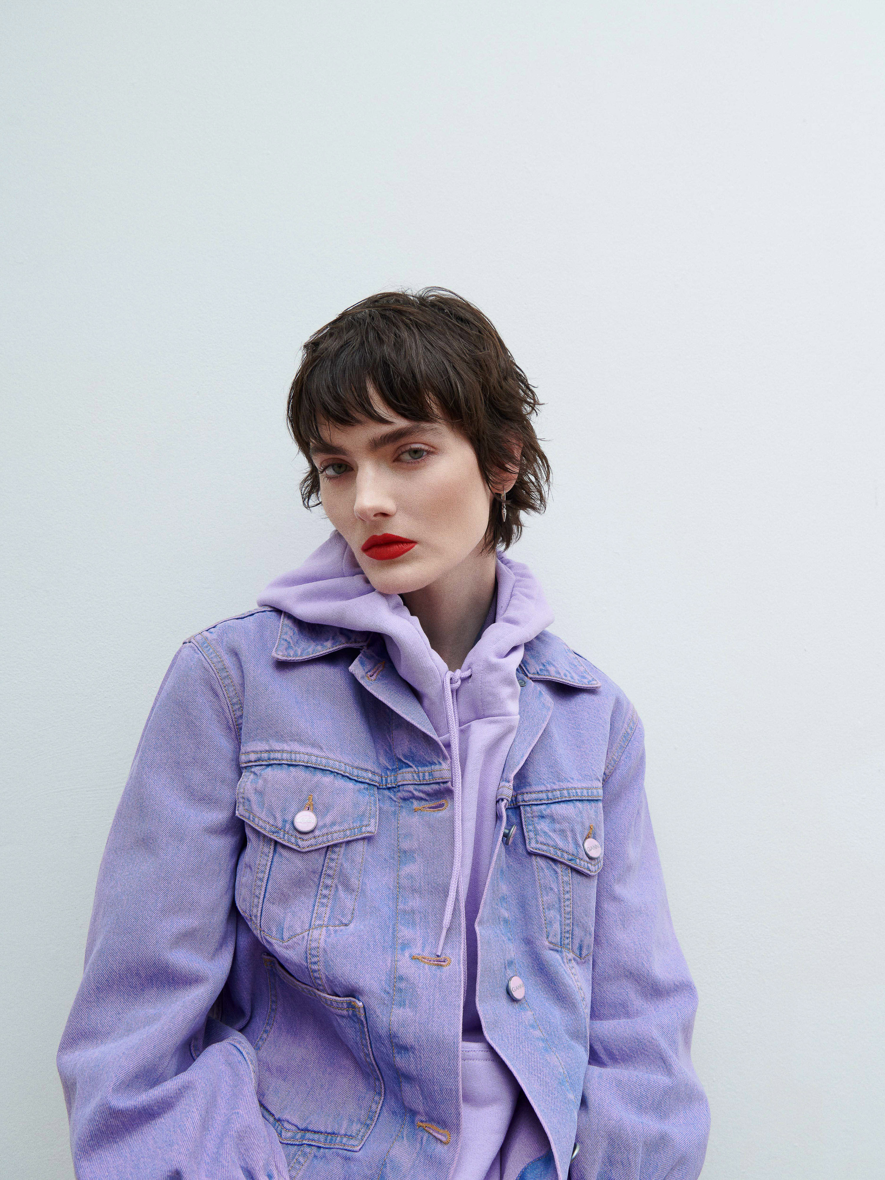 A fashionable woman in a lilac denim jacket and matching hoodie strikes a pose against a white backdrop, showcasing vibrant red lipstick.
