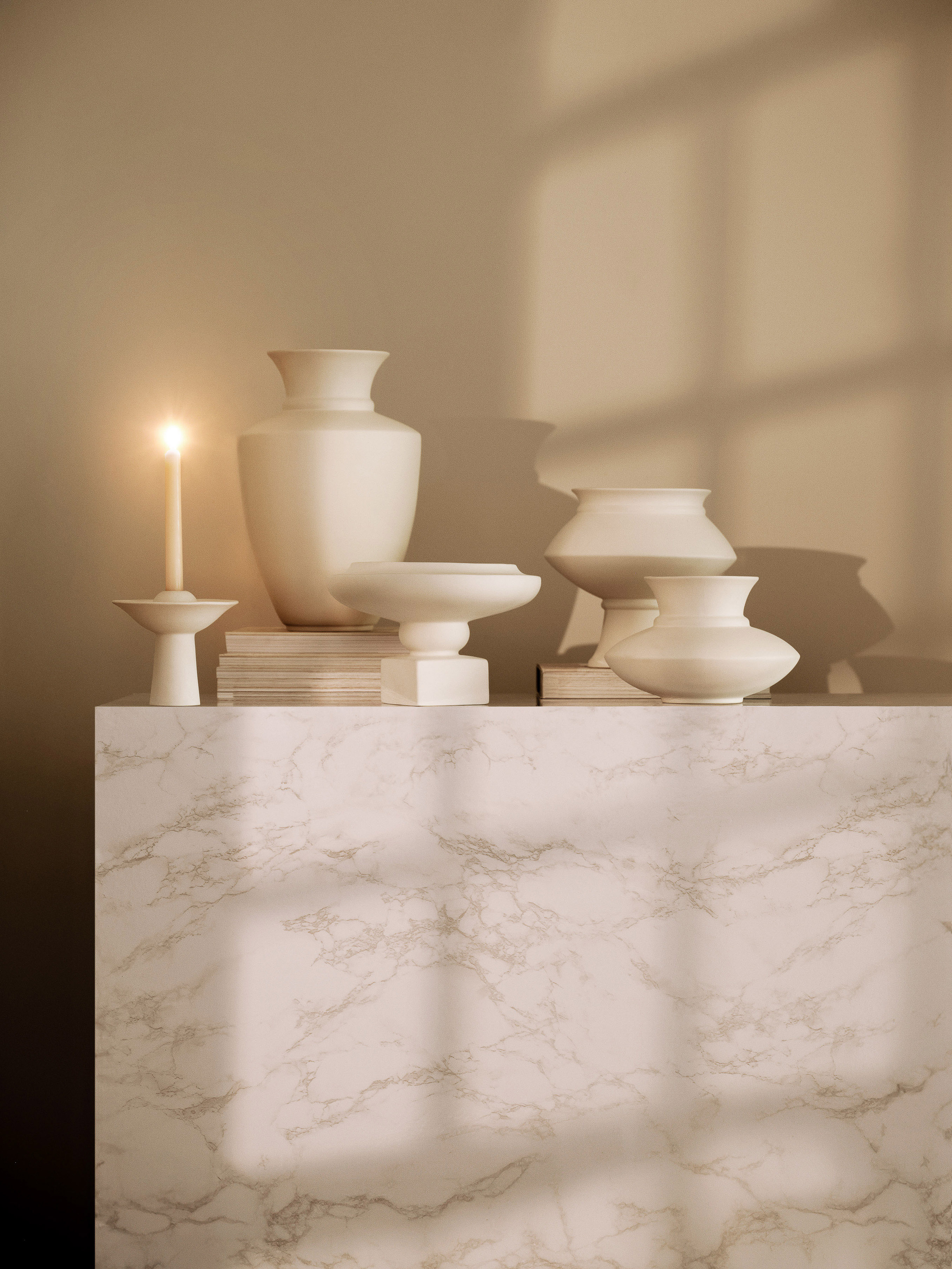 Still life with vases, books, and a candle on a marble surface, featuring warm lighting and shadows in a domestic setting.