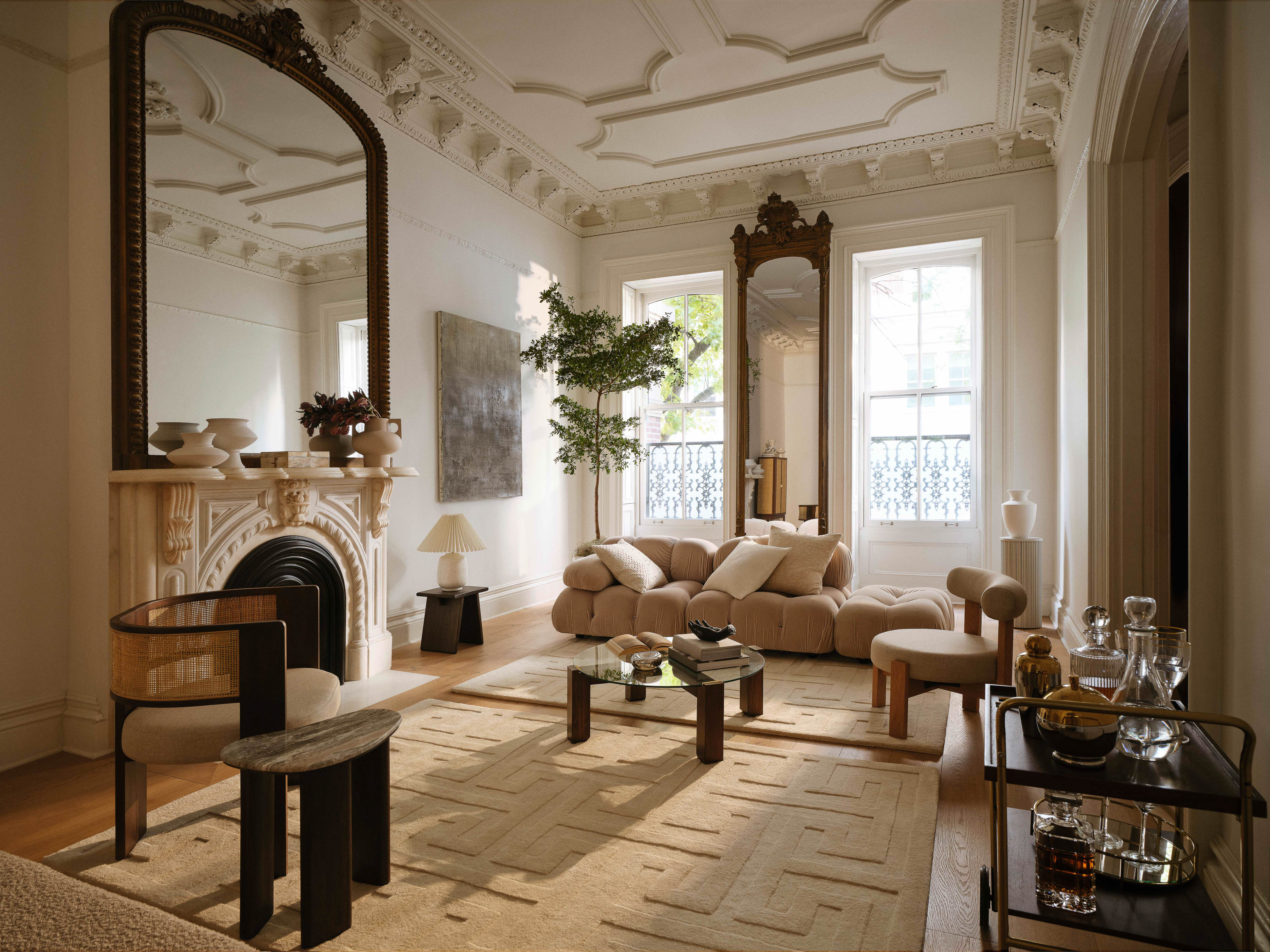 Living room interior with a beige sectional sofa, textured rug, coffee table, mirrors, fireplace, and decorative objects creates a cozy atmosphere.