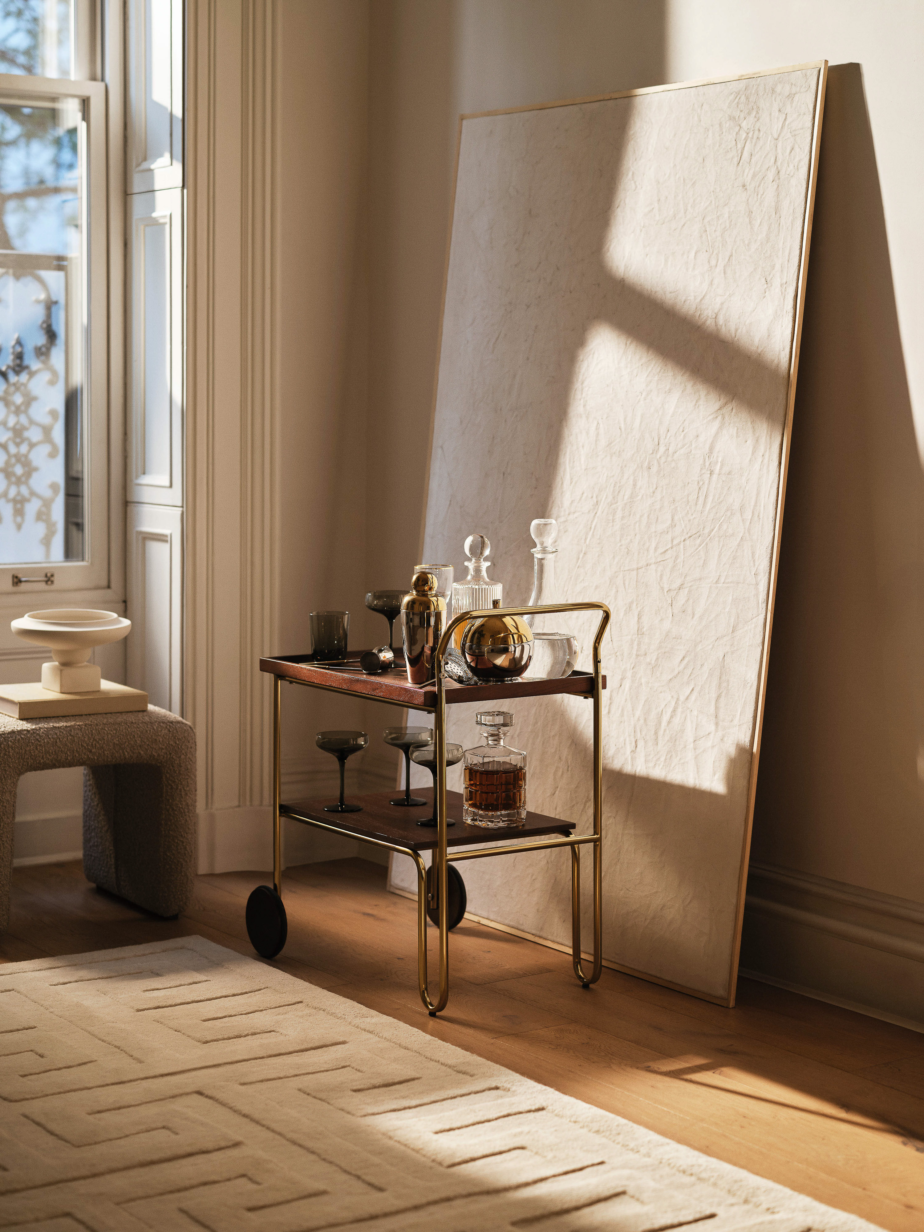 A rolling bar cart featuring glassware sits indoors on a patterned rug in front of a blank canvas and a window, casting shadows.