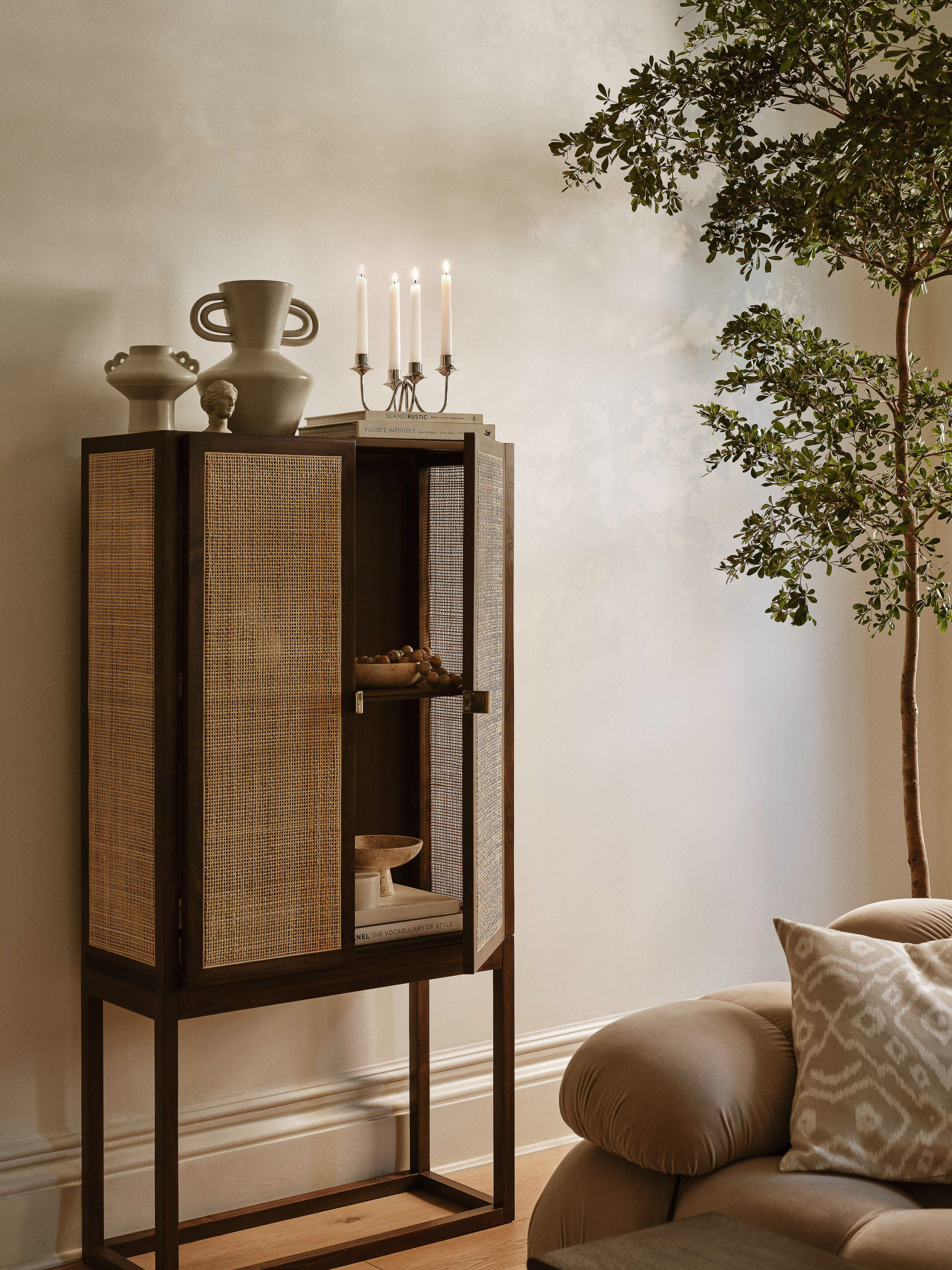 Rattan cabinet with pottery, candles, and books in a living room setting. Interior design featuring warm neutral tones and natural light.