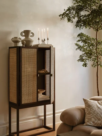Rattan cabinet with pottery, candles, and books in a living room setting. Interior design featuring warm neutral tones and natural light.