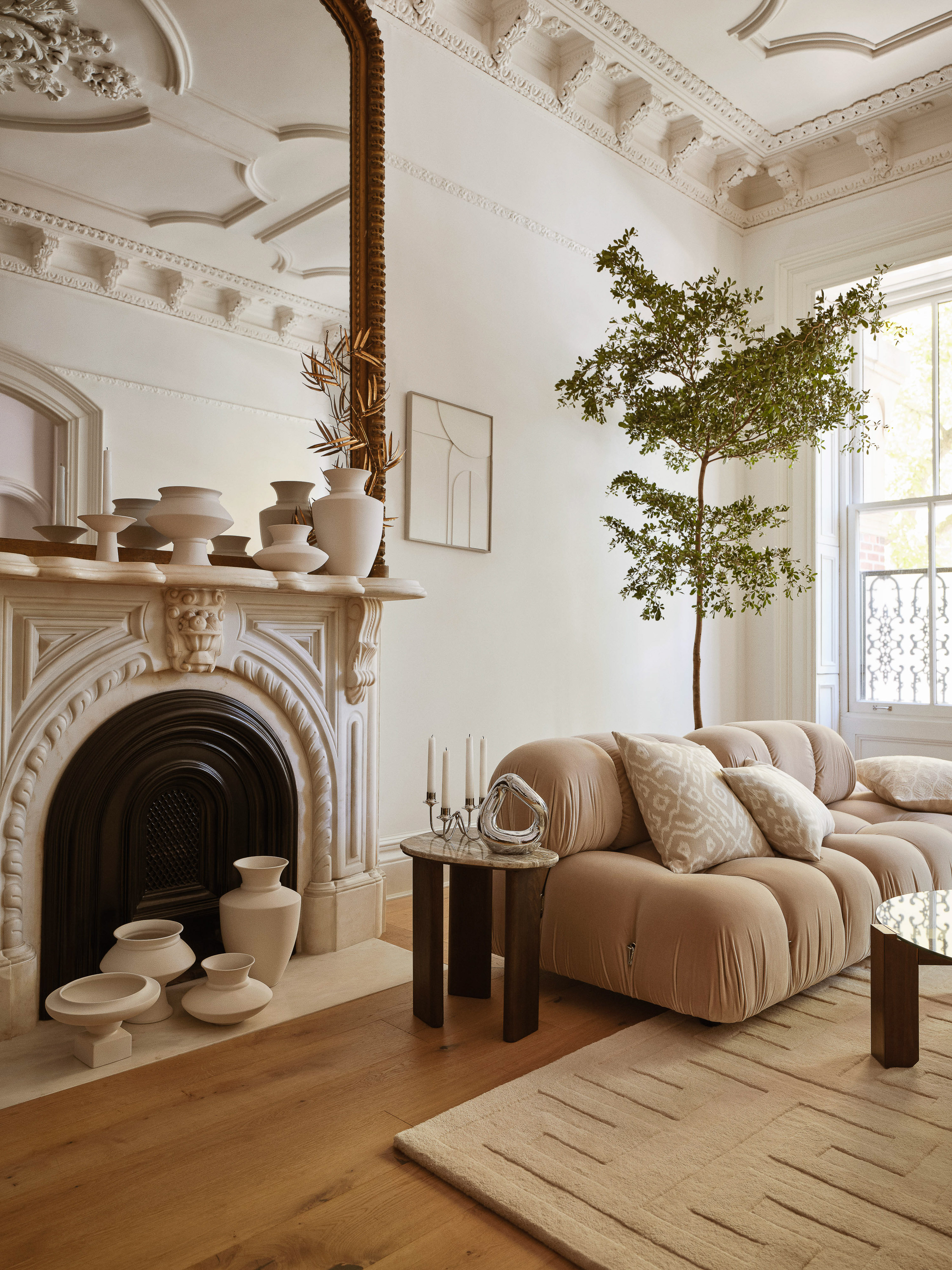 Beige sectional sofa in a living room with a fireplace, decorative vases, marble mantel, round side table, tree, and geometric rug.