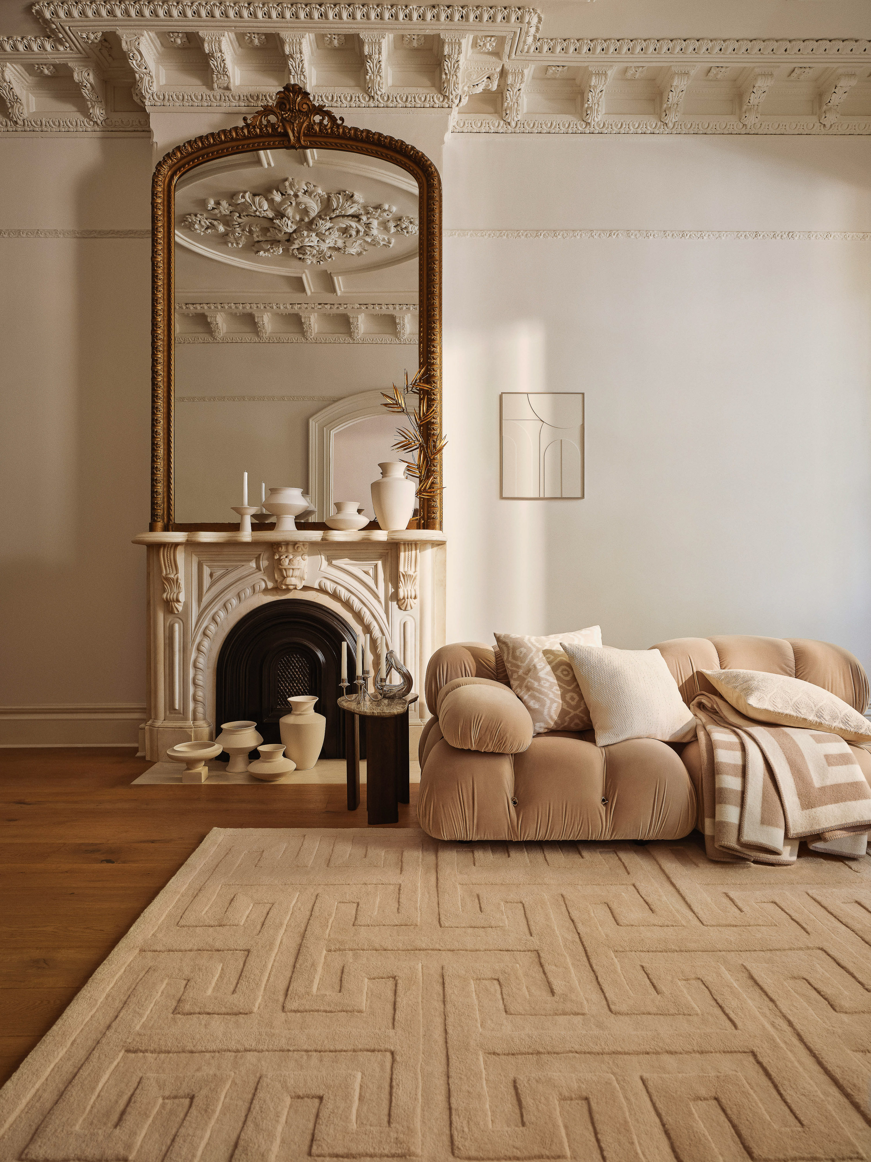 Living room interior featuring a neutral palette, patterned rug, couch with pillows, ornate mirror over fireplace, and decorative vases.