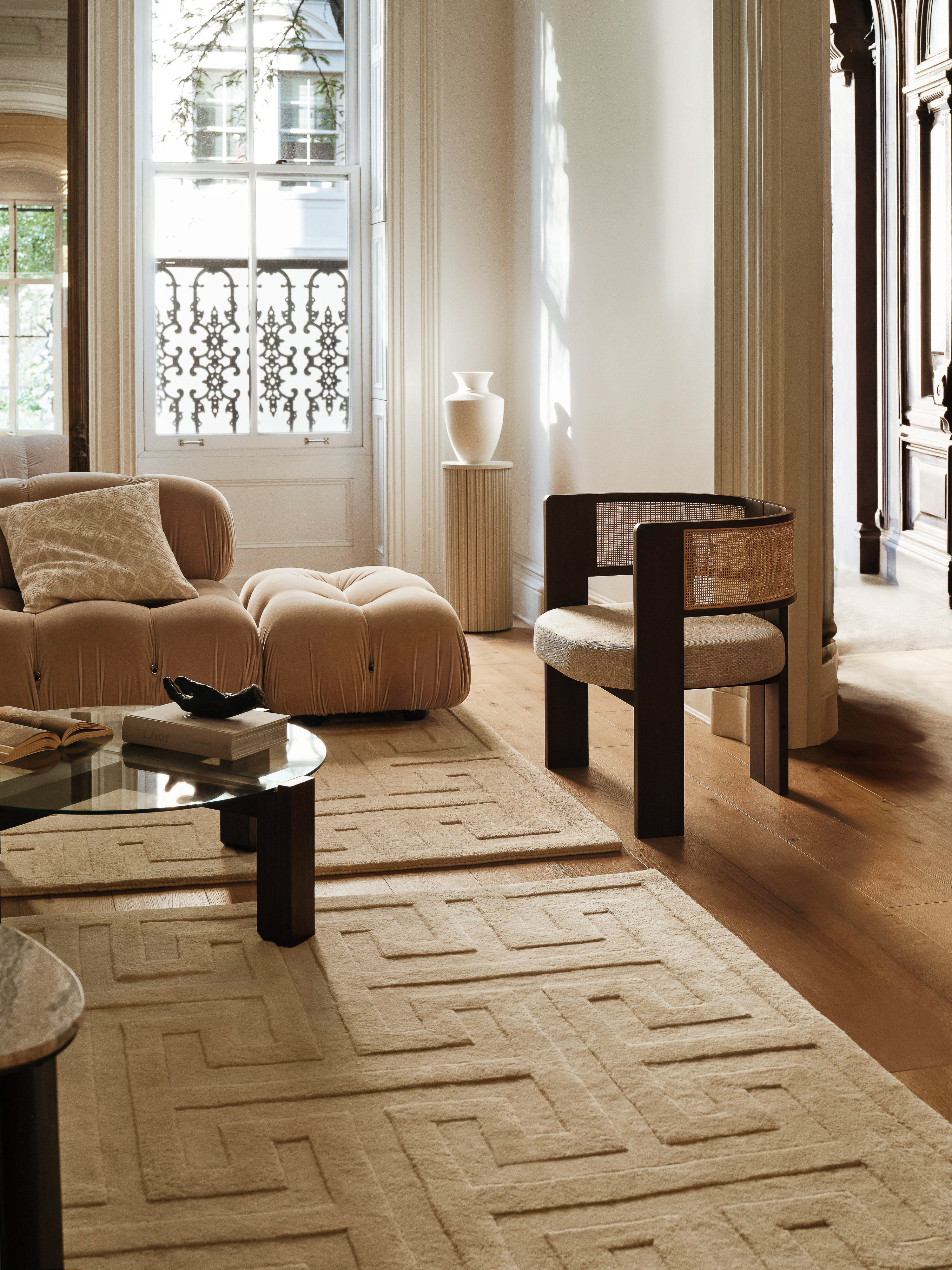 Interior design living room with tan colored sofa and ottoman, glass-topped coffee table, patterned rug, and modern armchair by window.