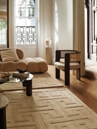 Sunlit living room interior with beige geometric patterned rugs, a tufted modular sofa, and a cane-back accent armchair.