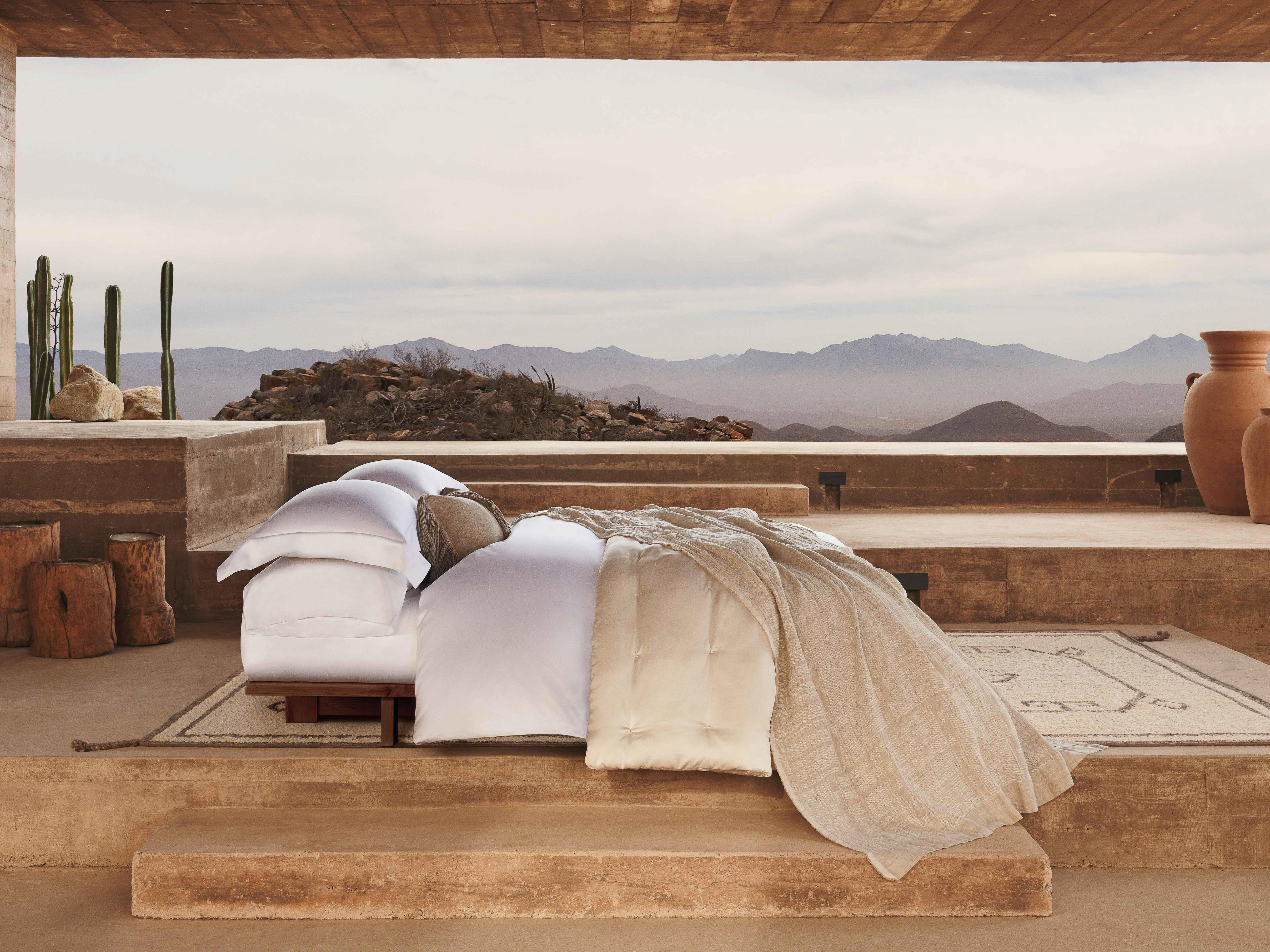 Outdoor bedroom setup with white bedding and a woven throw blanket, set against a landscape of mountains under an overcast sky.