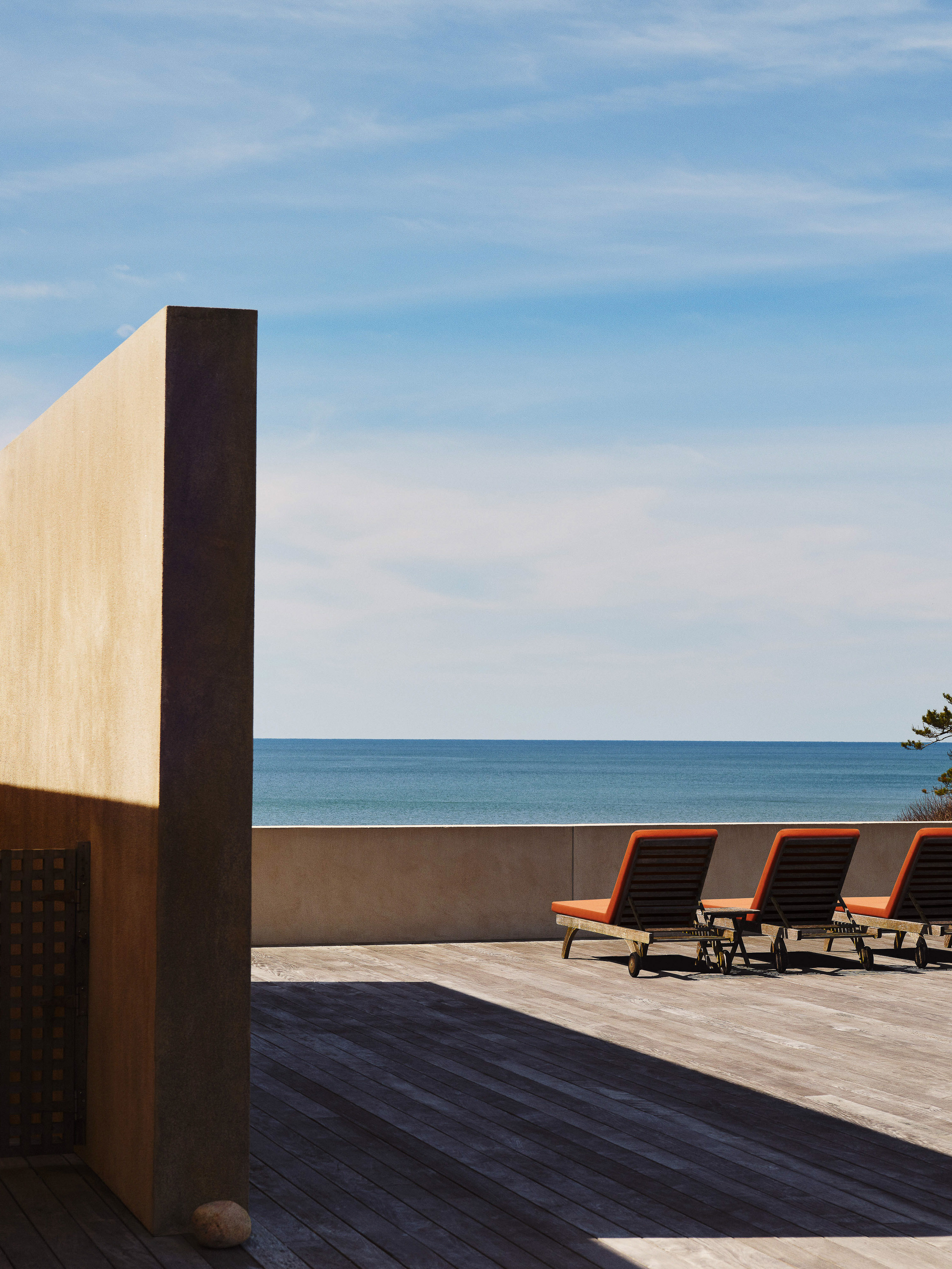 Minimalist outdoor architecture featuring orange lounge chairs on a wooden deck with ocean view under a blue sky in daytime.