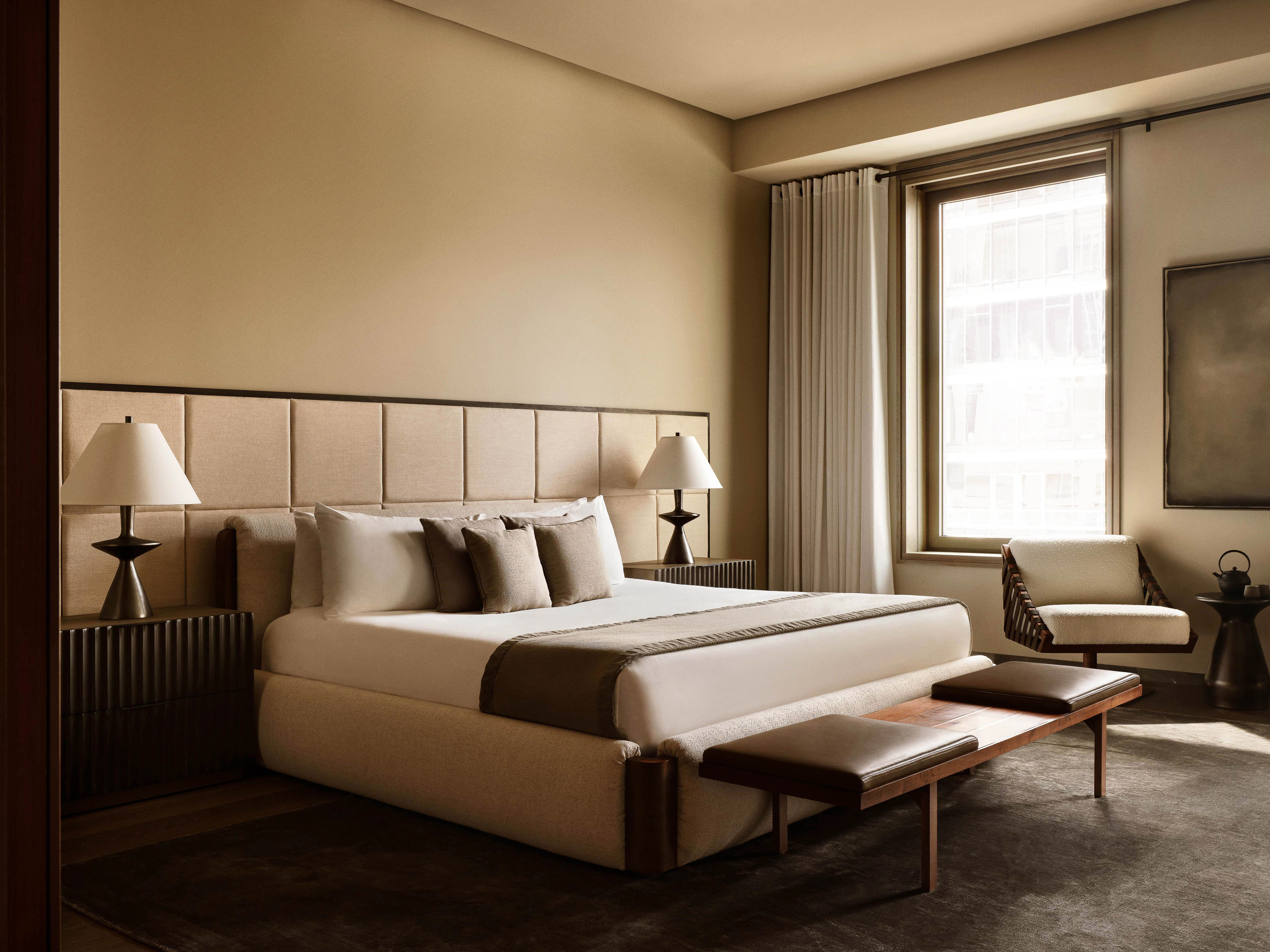 Neutral-toned bedroom interior with a large bed, padded headboard, bench, armchair, and window with natural light in a modern style.