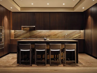 Modern luxury kitchen featuring dark wood cabinetry, an earth-toned stone backsplash, and four bar stools at a central island.