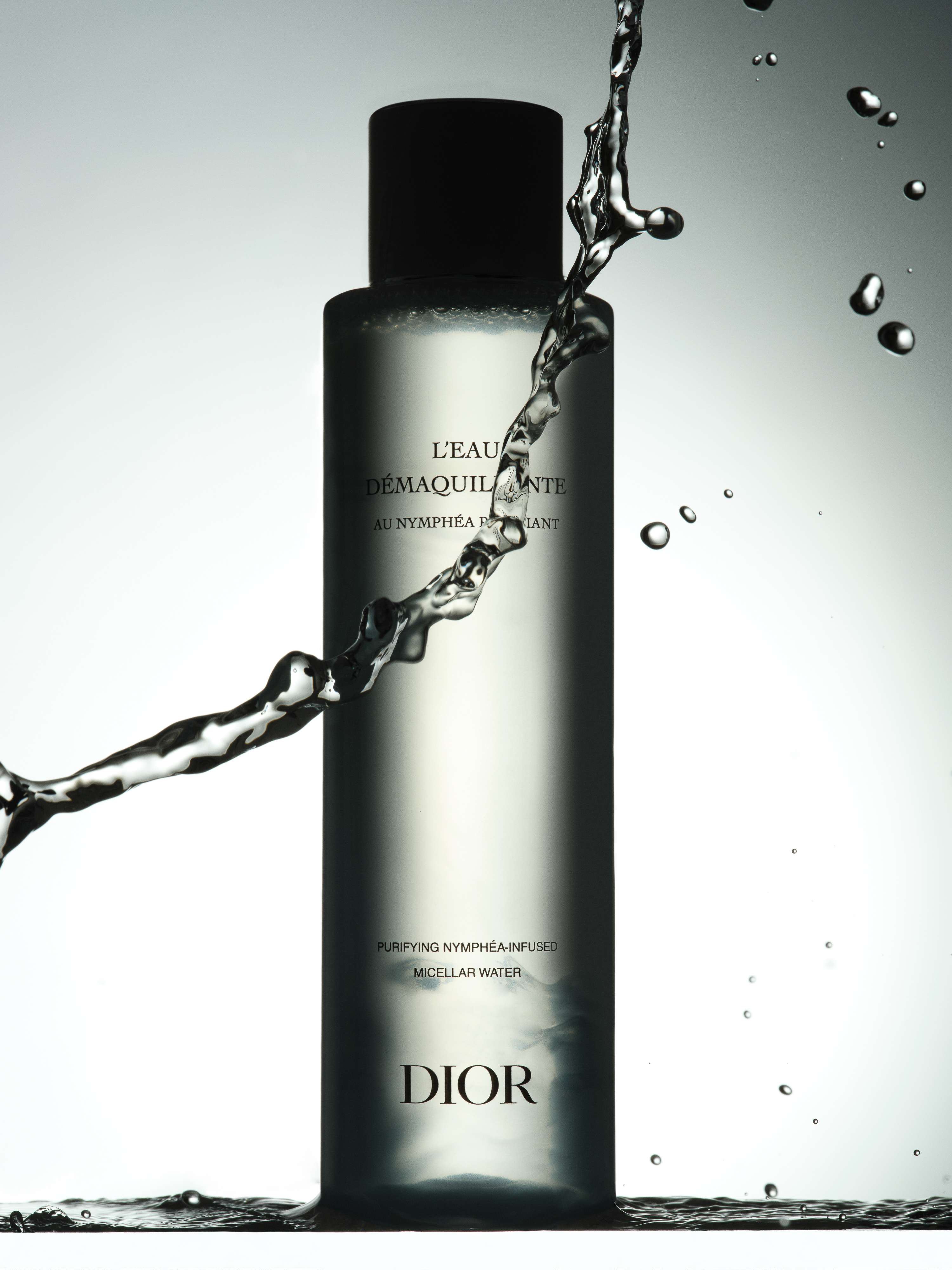 Cosmetic bottle with water splash. Clear glass bottle with black cap and Dior logo against a light background. Beauty shot.