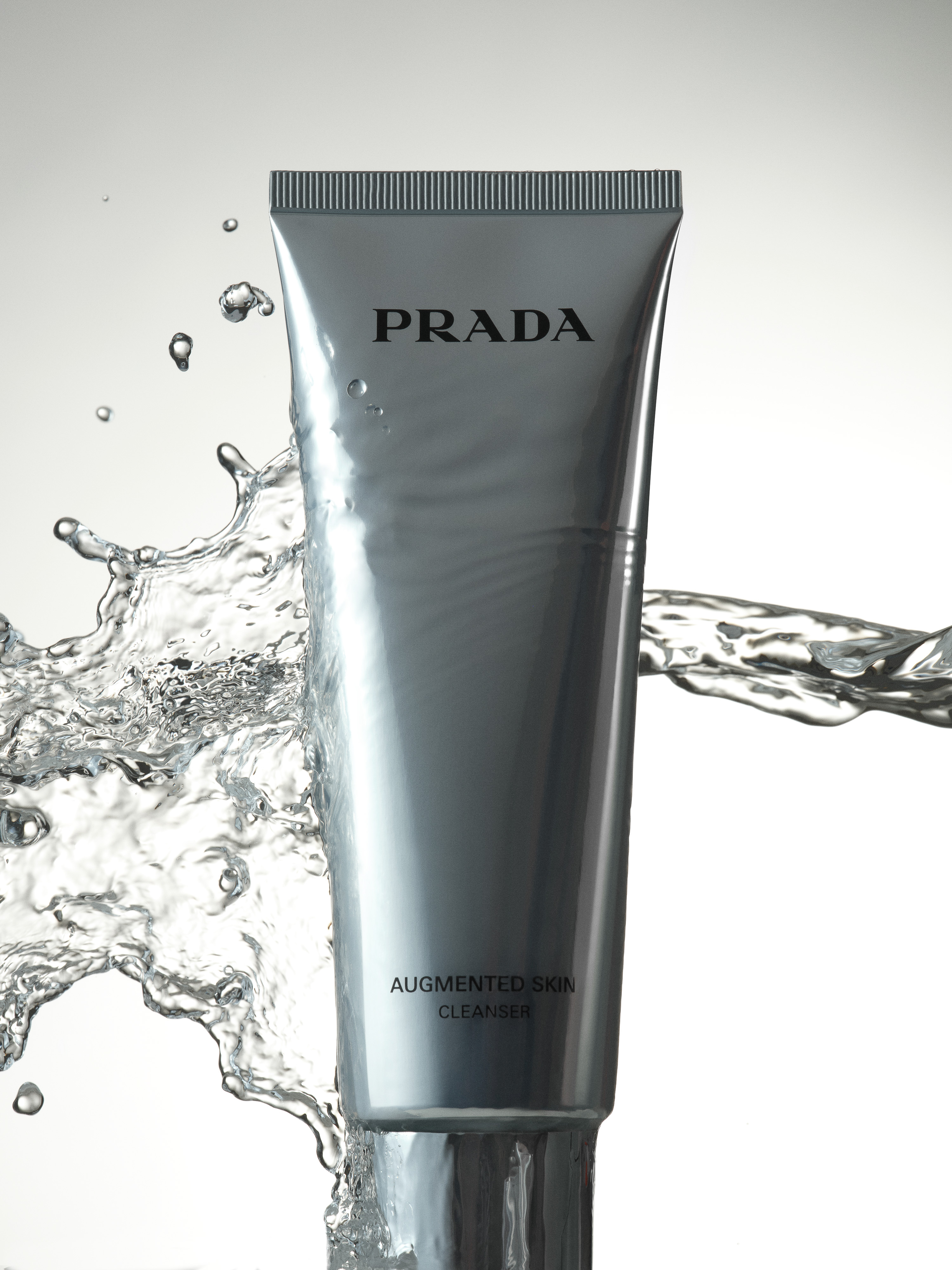 A grey Prada Augmented Skin Cleanser tube is surrounded by splashing water on a white background in a beauty product shot.