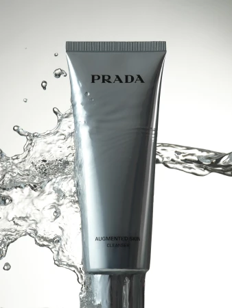 A grey Prada Augmented Skin Cleanser tube is surrounded by splashing water on a white background in a beauty product shot.