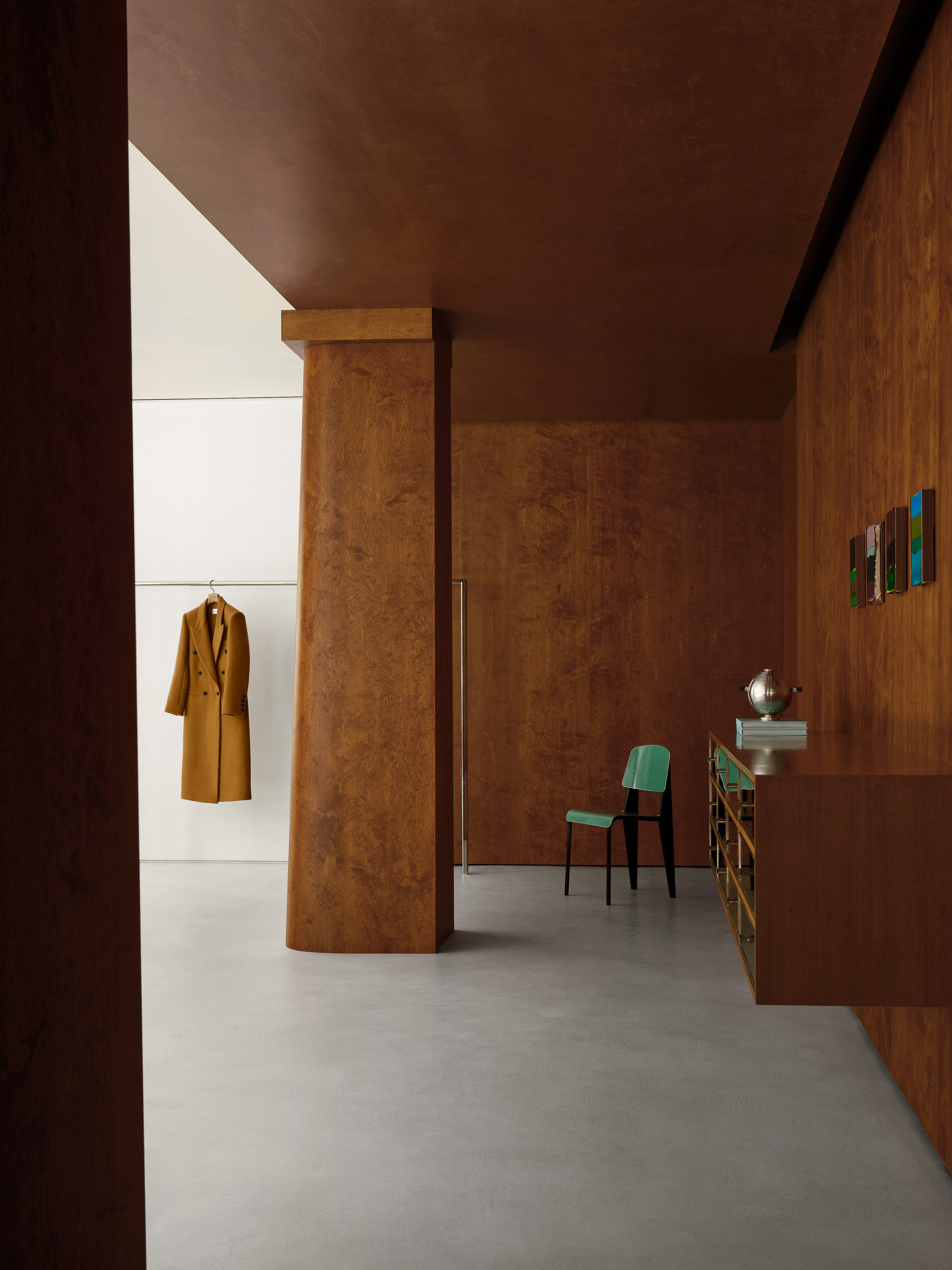Interior design featuring a tan coat hanging on a white wall, a green chair, brown furniture, and brown walls in a minimalist setting.