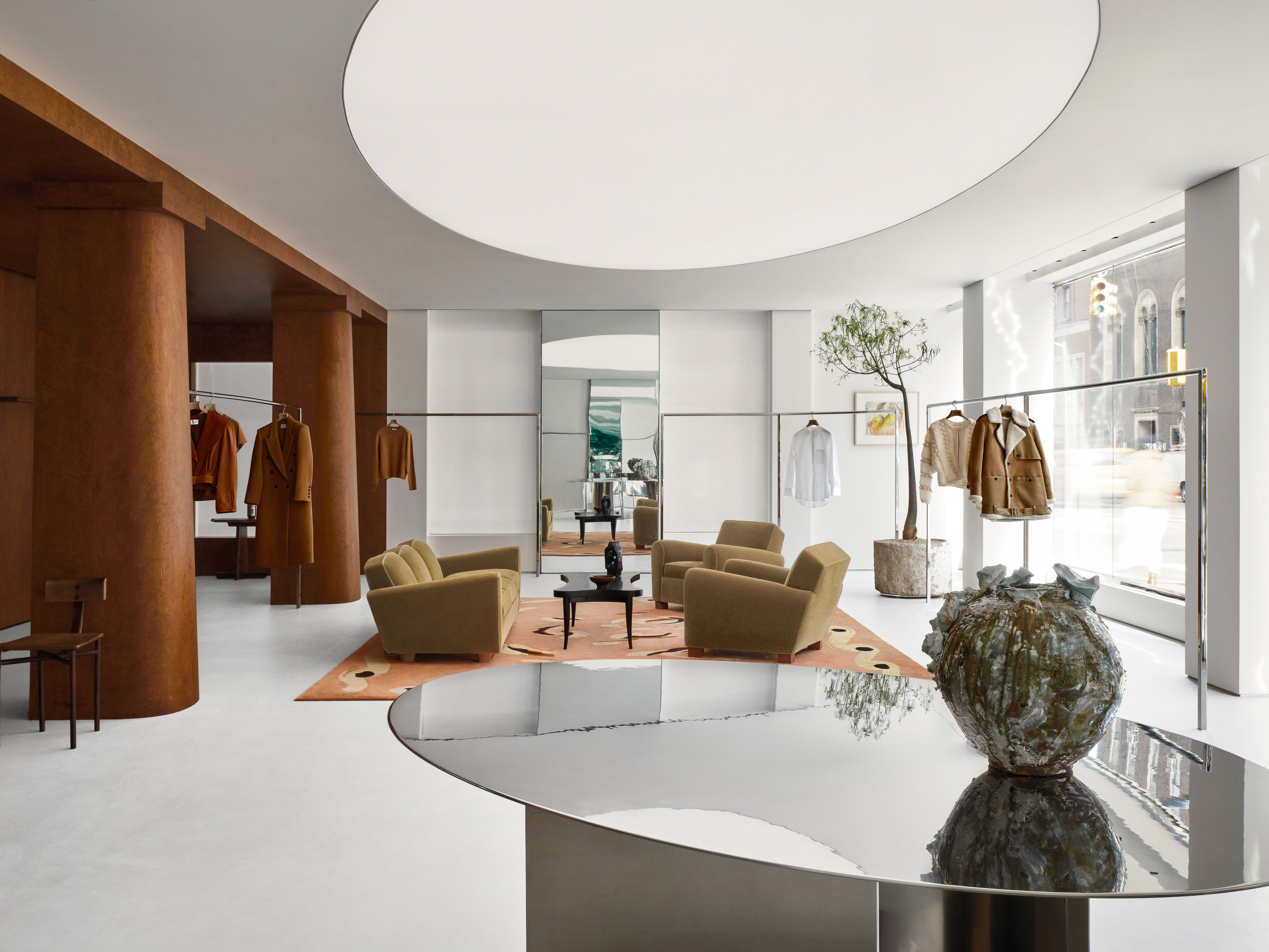 Interior view of a fashion boutique featuring clothing racks with apparel, seating areas with sofas, and a reflective display table in the foreground.