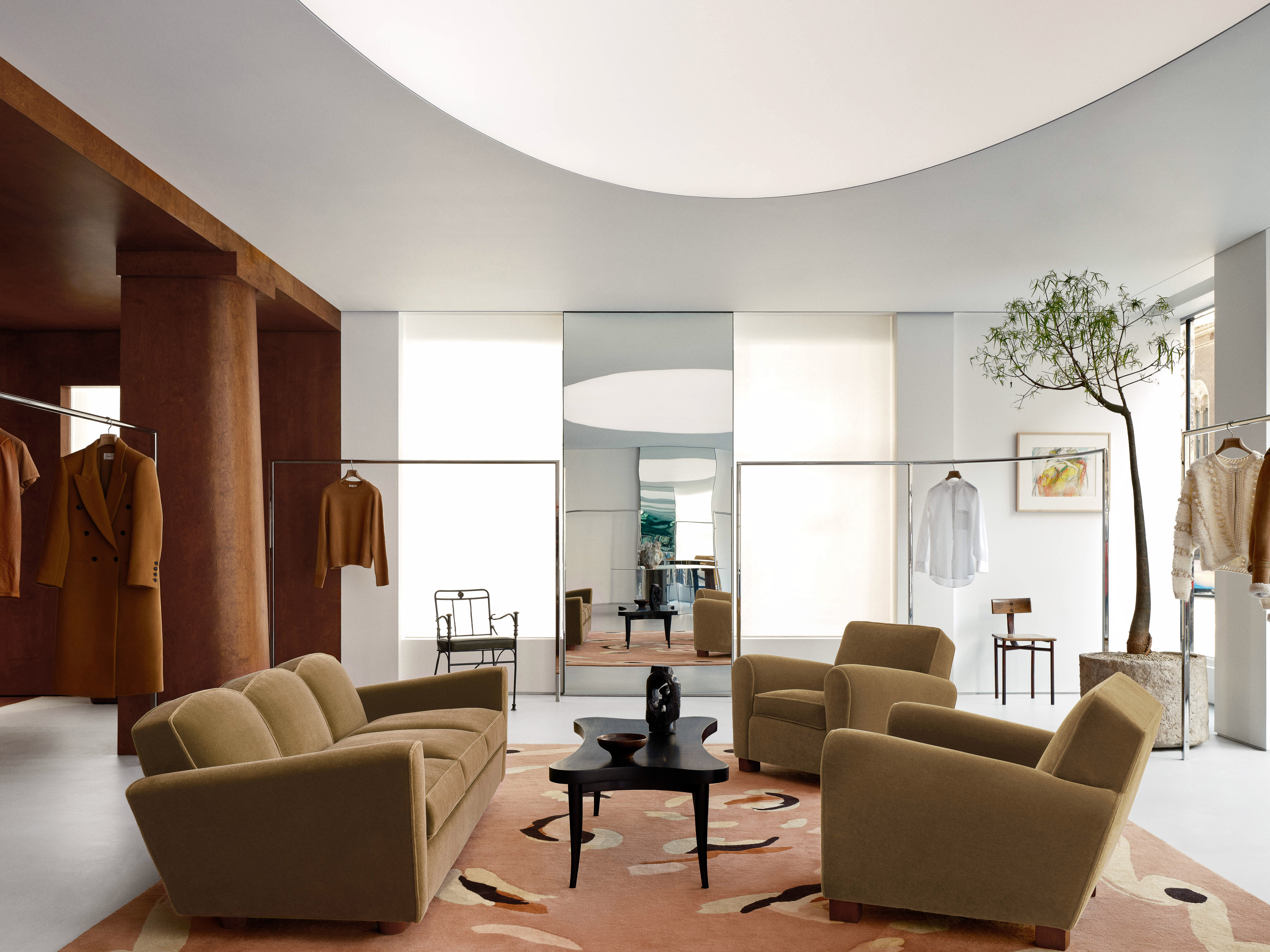 Living room interior with tan sofa, armchairs, patterned rug, dark wood table, clothing racks, white walls, brown column, and plant.