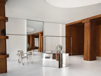 Minimalist boutique interior featuring a mirrored metallic display table, sculptural chair, and warm wooden architectural columns.