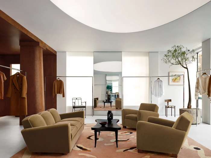 Spacious minimalist fashion store interior with velvet lounge seating, patterned rug, clothing racks, and an indoor potted tree.