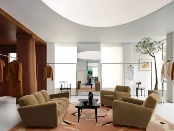 Spacious minimalist fashion store interior with velvet lounge seating, patterned rug, clothing racks, and an indoor potted tree.