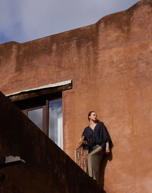Adult woman in a black top and tan skirt stands on a balcony with an old, stucco building with window and blue sky behind her.