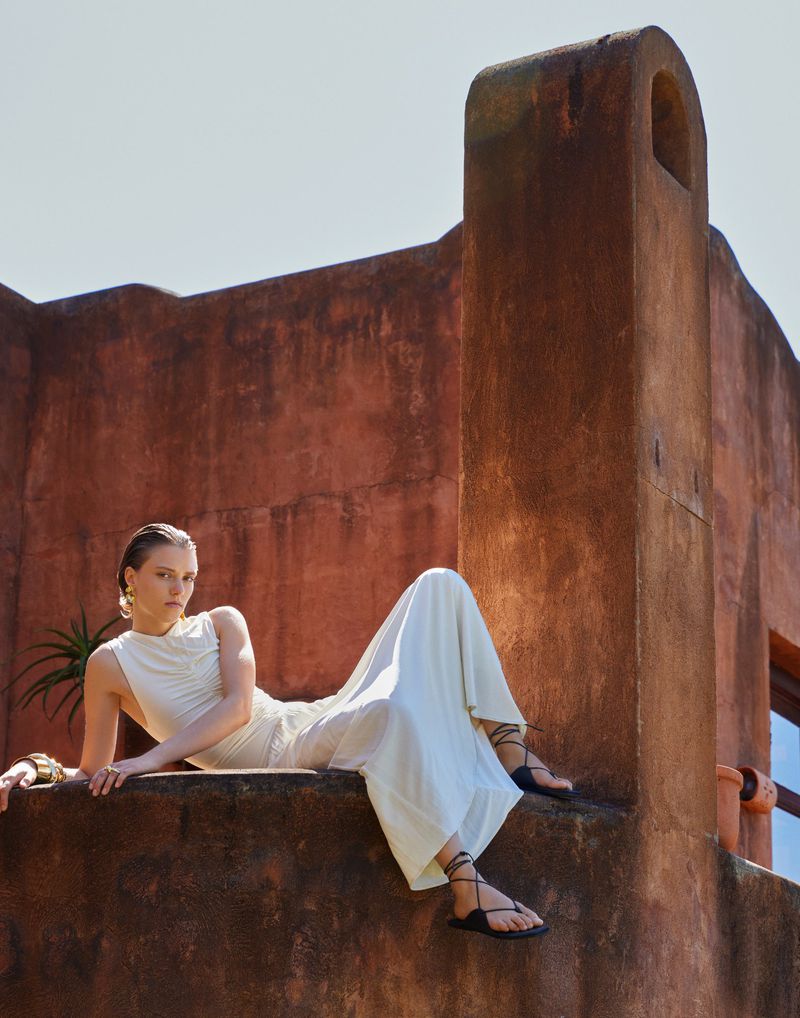 Woman in a white dress and sandals reclines on a textured wall under bright sunlight; terracotta building in the background.