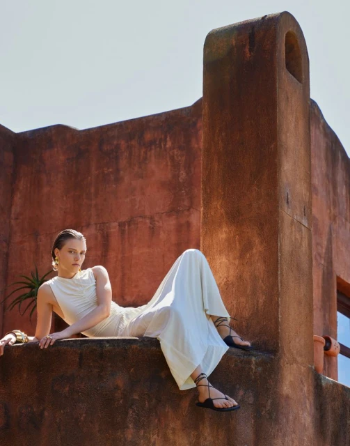 Woman in a white dress and sandals reclines on a textured wall under bright sunlight; terracotta building in the background.