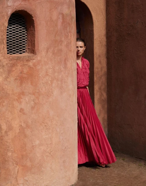 Adult female in red dress stands in an archway with a textured wall in the background and window with mesh covering.