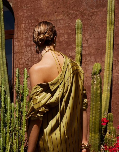 Back view of an adult female in a striped olive-green outfit, accessorized with a gold bracelet, posing in front of a brick wall and cacti.