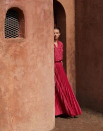 Model in a bright pink pleated dress peeking around a textured terracotta architectural wall