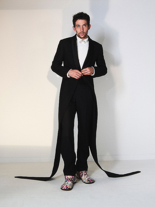 Full-length studio shot of a man in a black tuxedo jacket, white shirt, and black pants with long black fabric strips attached, striking a pose.