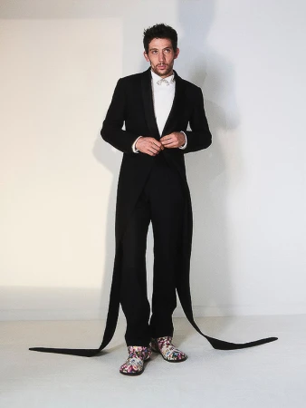 Full-length studio shot of a man in a black tuxedo jacket, white shirt, and black pants with long black fabric strips attached, striking a pose.