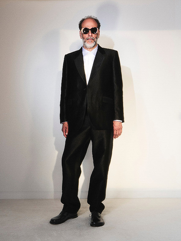 Man in a black velvet suit, white shirt, and sunglasses posing against a light wall in a full-body shot with a neutral expression.