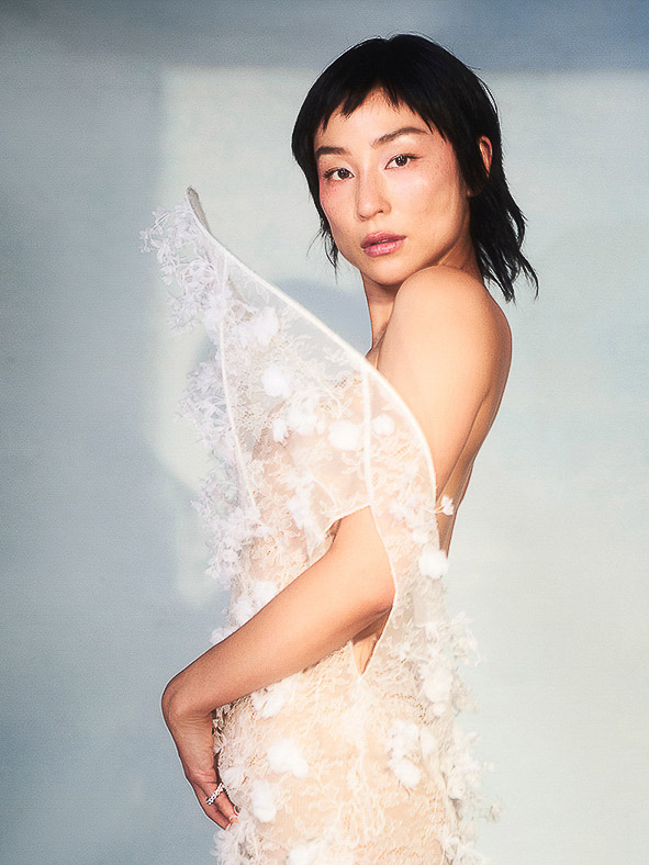 Portrait of a young Asian woman in a white, floral lace gown, posing in front of a soft blue background in a studio setting.