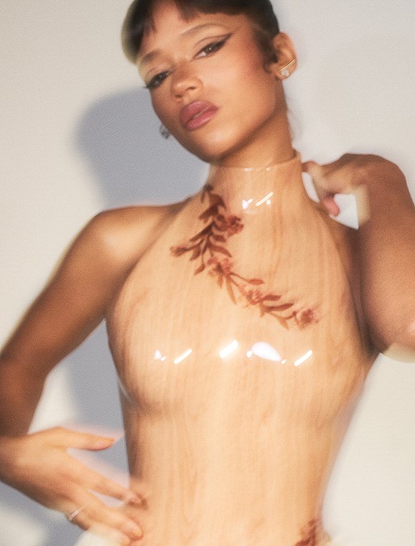 Elegant woman in a tan halter dress with floral accents, posing against a soft white background in a fashion-forward, studio shot.
