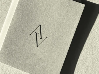 Textured off-white paper card printed with an intertwined black serif AV monogram logo casting a shadow on textured paper.