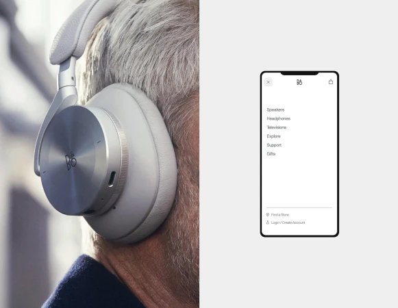 Close-up of silver Bang & Olufsen headphones worn by a person with gray hair; out-of-focus mobile phone displays B&O interface.