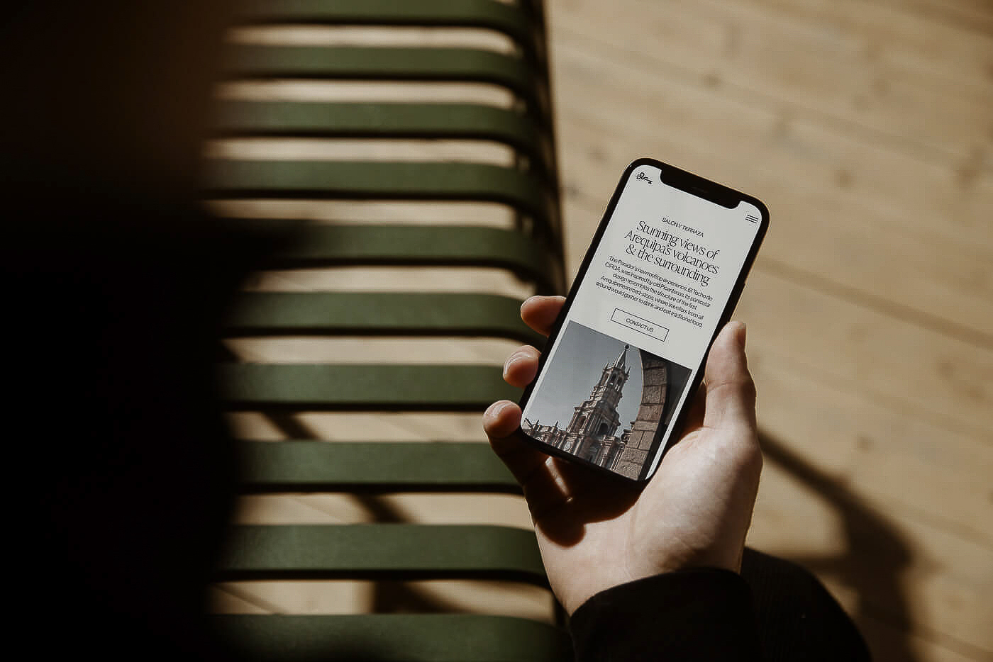 Person holds a smartphone displaying a travel website featuring a cathedral, with a bench and sunlight in the background.