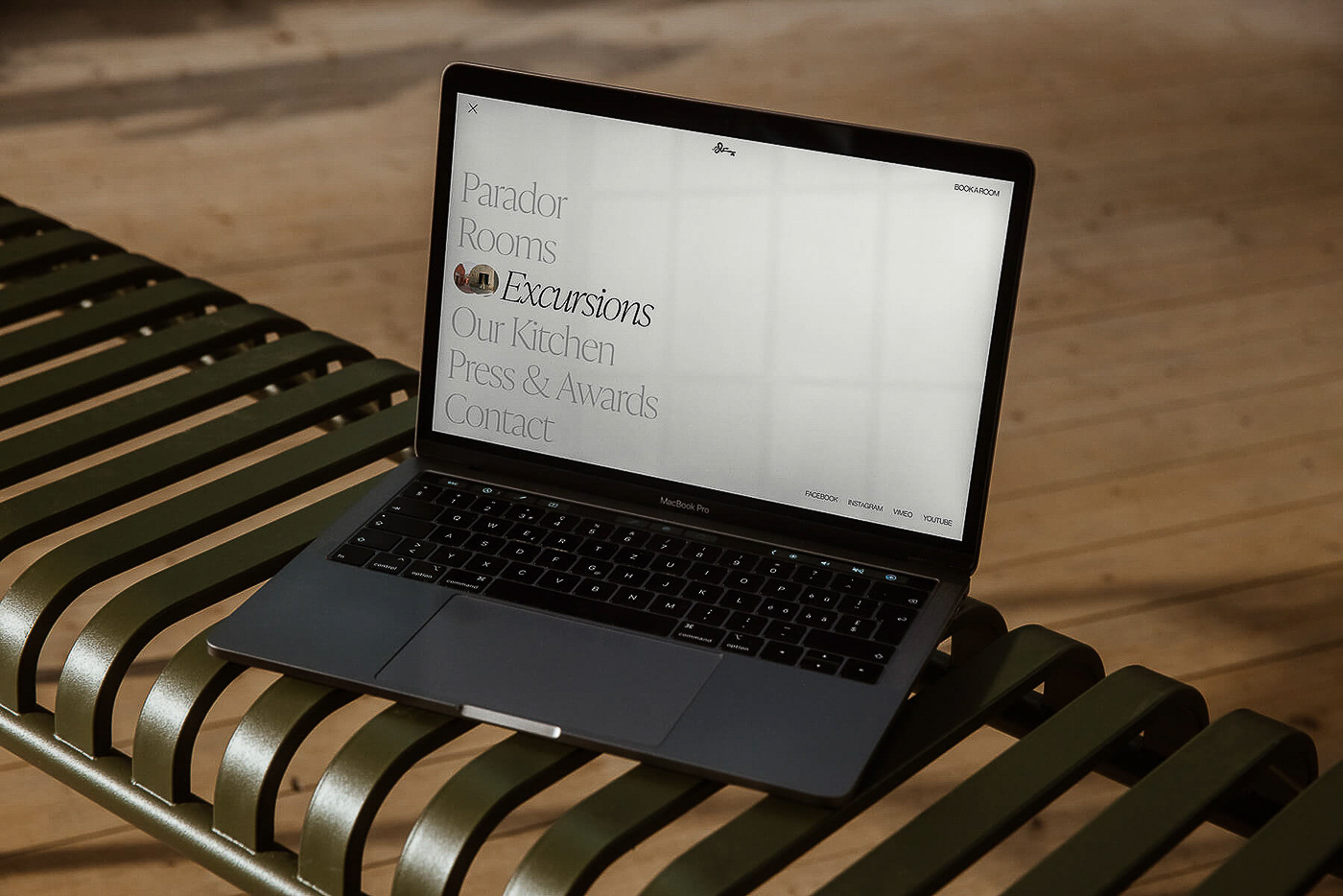 Laptop on a bench displays a website with text 'Parador, Rooms, Excursions, Our Kitchen, Press & Awards, Contact'.