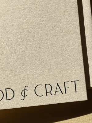 Close-up of Food & Craft branding printed in black serif typography on textured cream paper with sunlight casting a diagonal shadow.
