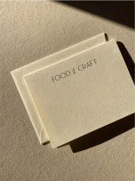 Cream textured stationery card and matching envelope printed with FOOD & CRAFT casting sharp diagonal shadows on paper.